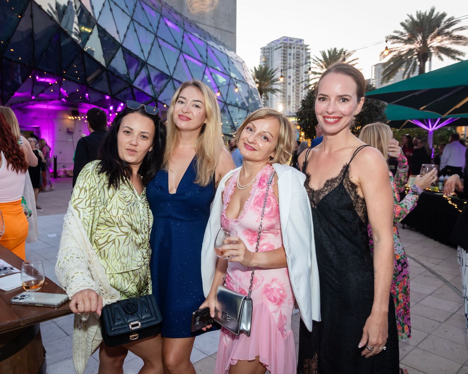 Four women posing together at an elegant outdoor evening event with colorful lights and palm trees.