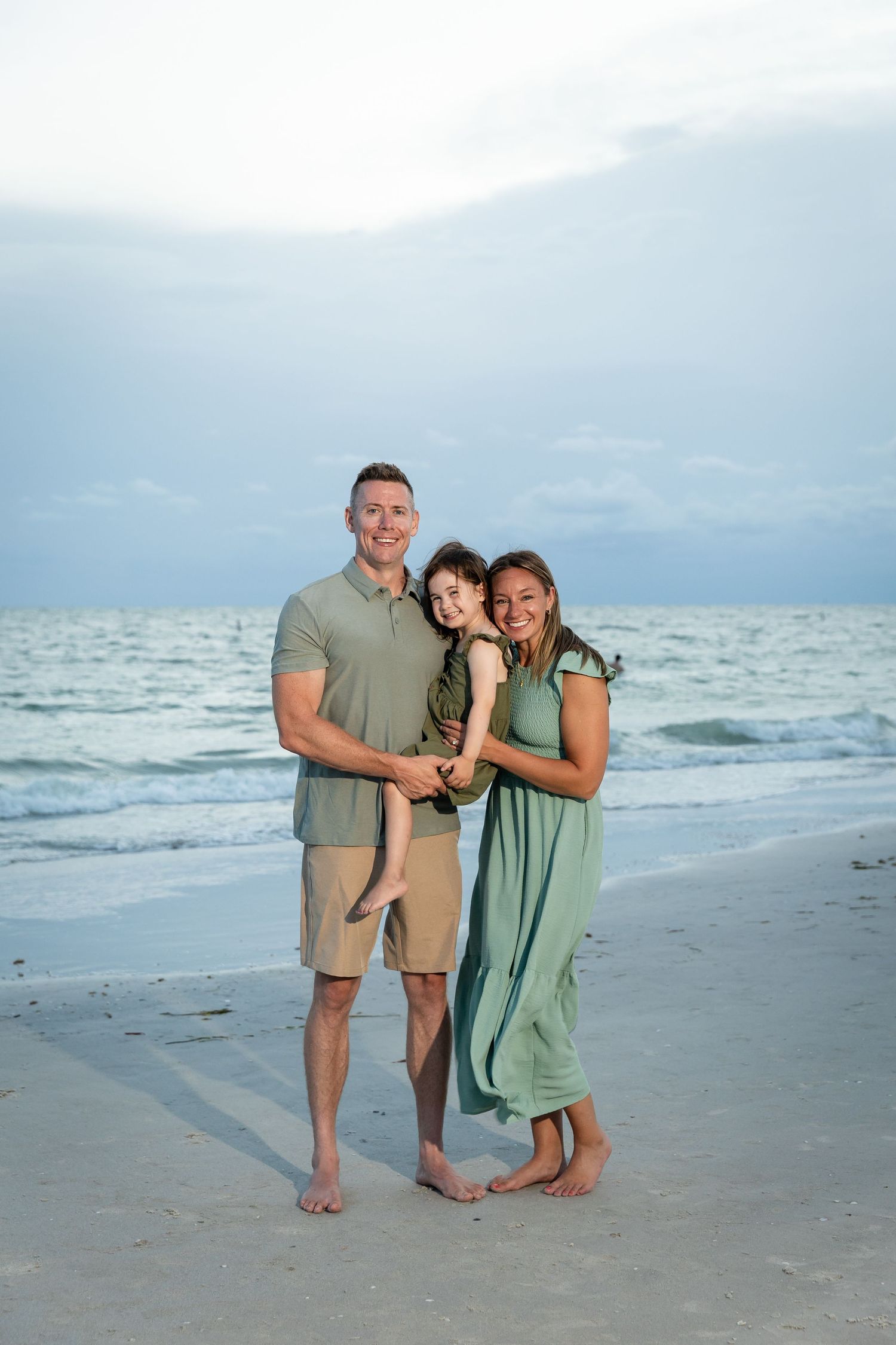 A happy family of three poses barefoot on a sandy beach at dusk, wearing coordinating green outfits.