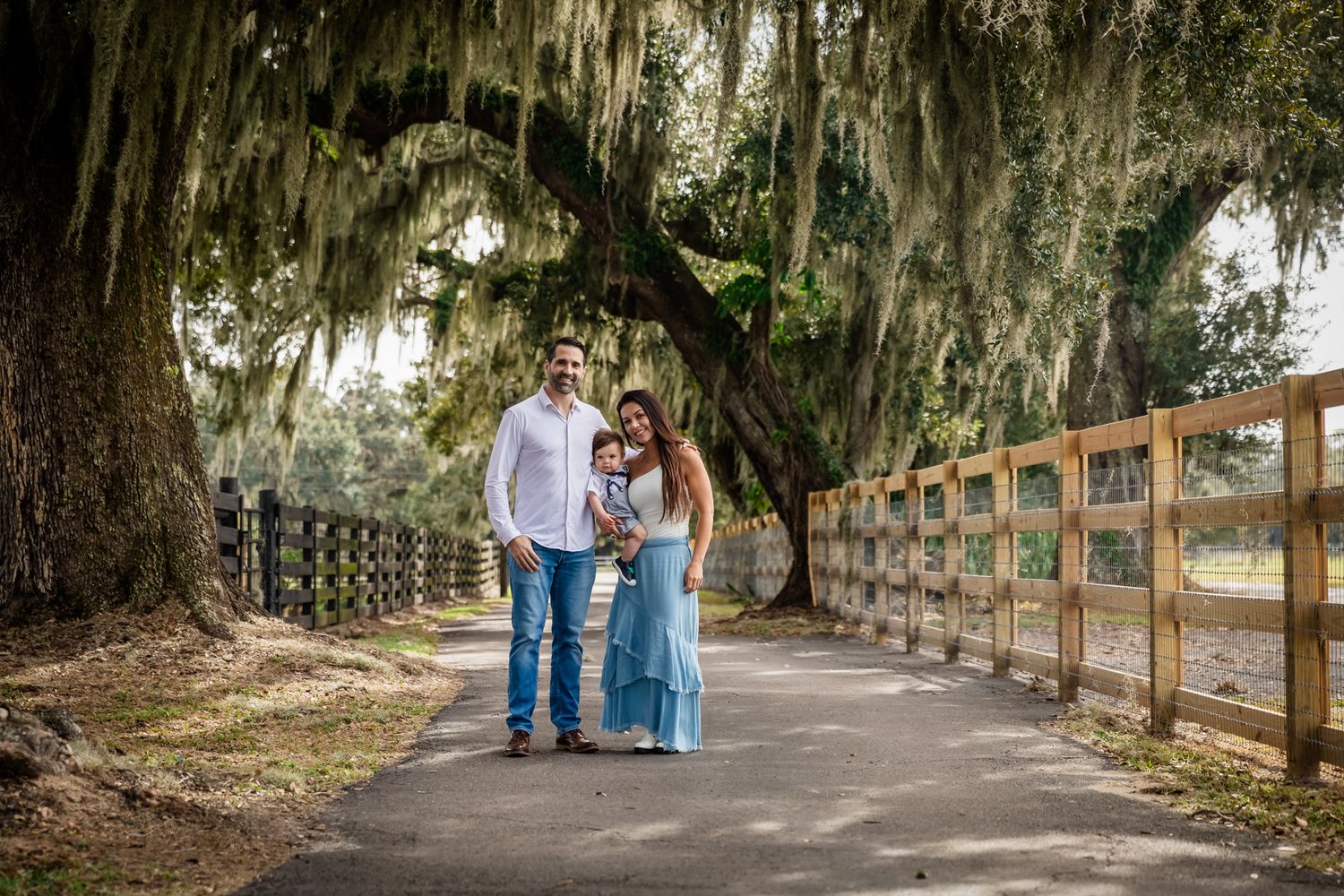 Family of three standing on tree-lined path with Spanish moss and wooden fence in background.