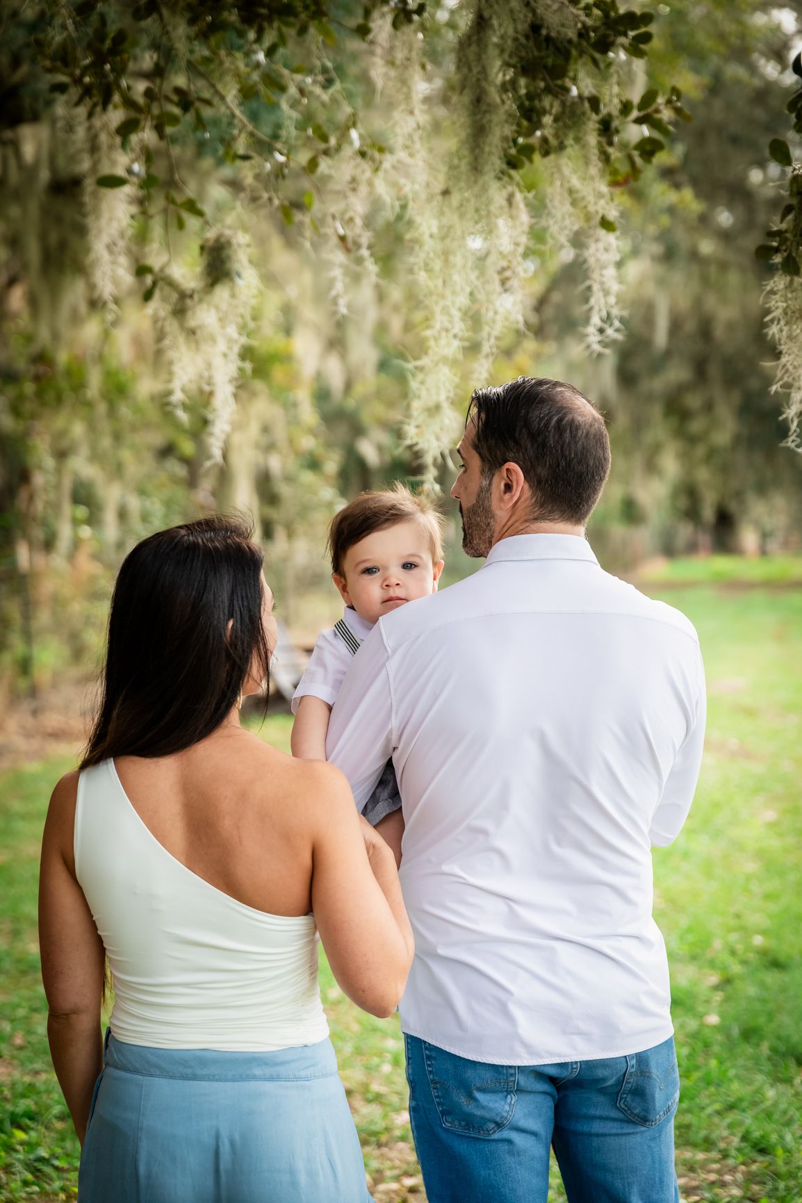 A family of three poses outdoors under hanging moss, parents holding their baby boy in a lush green park setting.