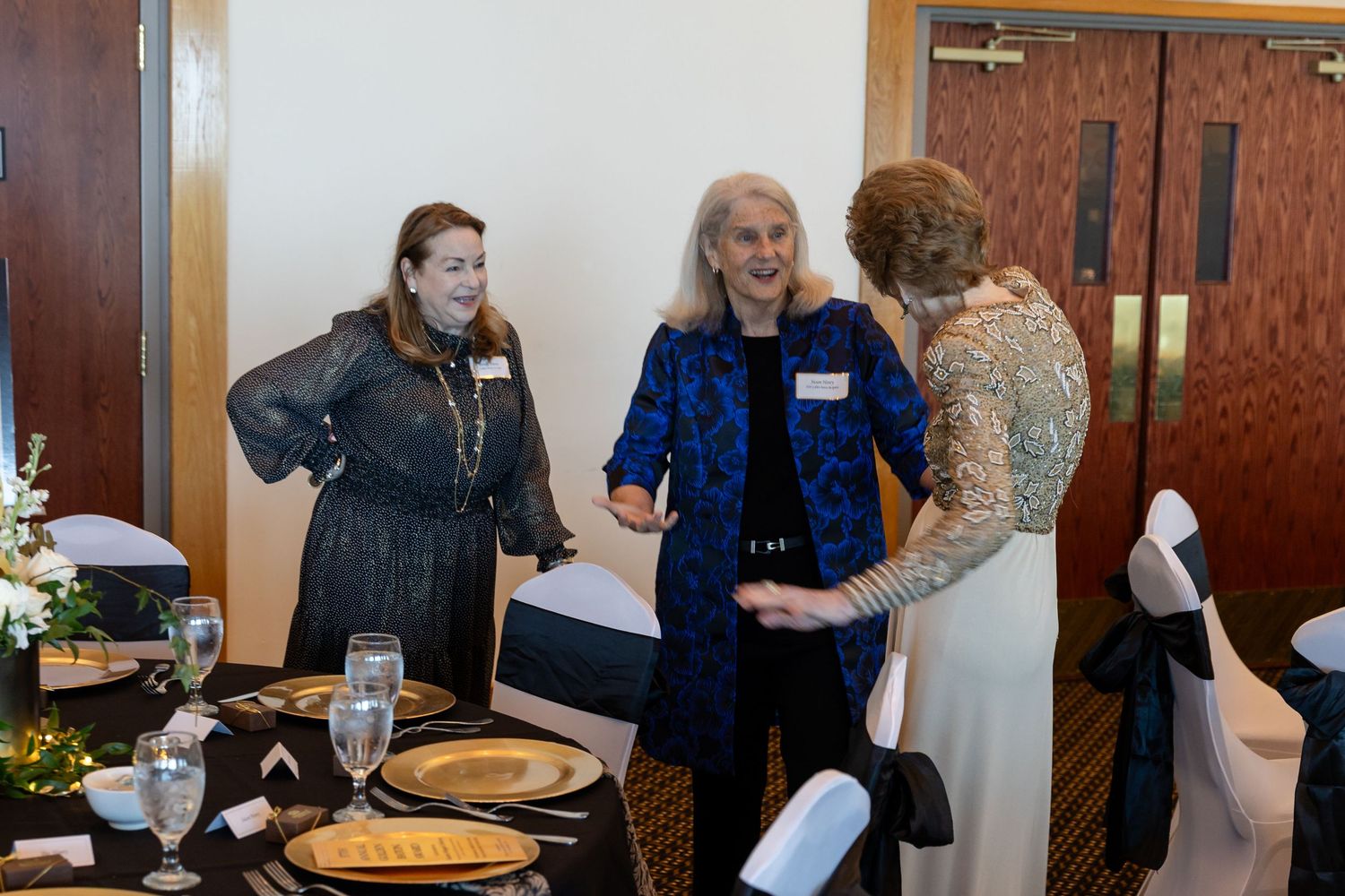 Three elegantly dressed women socializing at a formal banquet table set with gold plates and floral centerpieces.