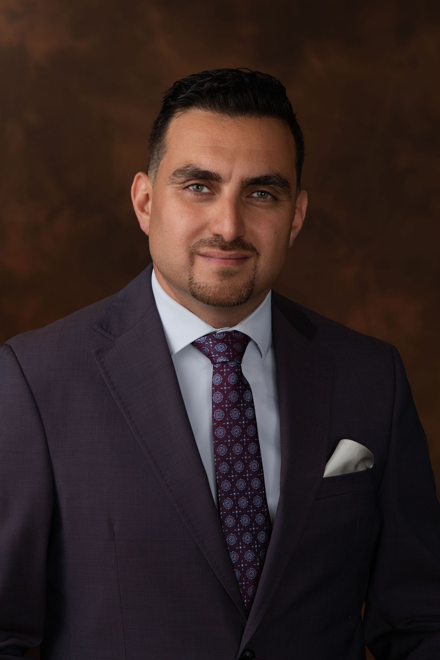 Professional man in dark suit and patterned tie poses for formal business portrait against brown background.