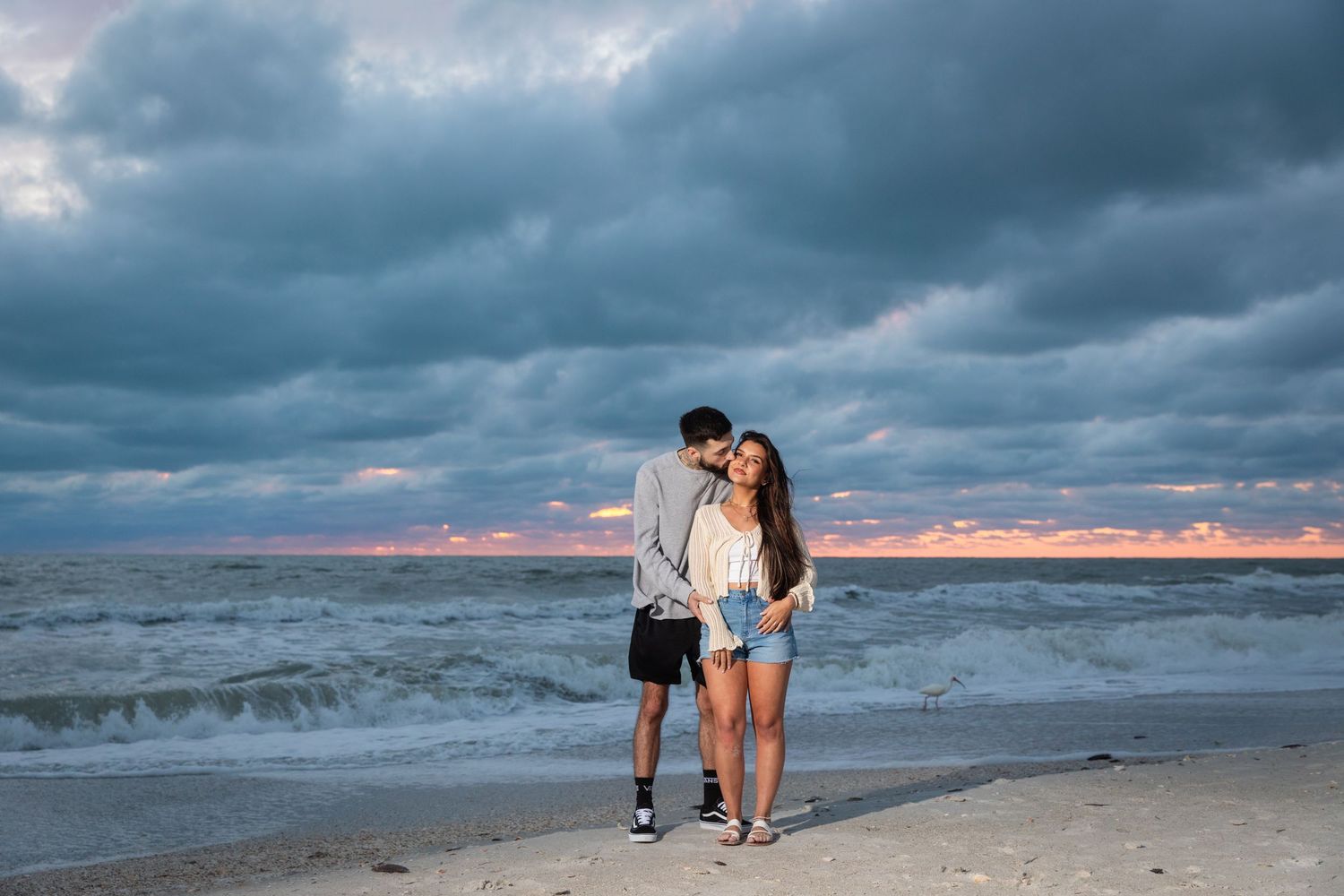 Couple embracing on a sandy beach at sunset with dramatic cloudy skies and ocean waves behind them.