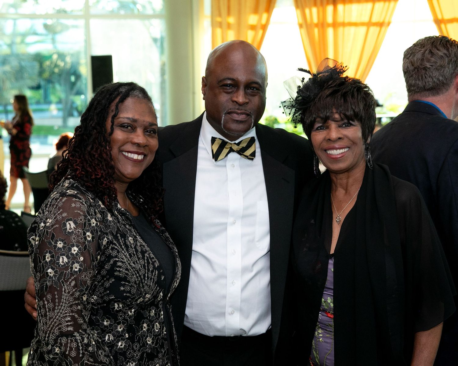 Three elegantly dressed people posing together at a formal event, man in tuxedo with bow tie flanked by two women.