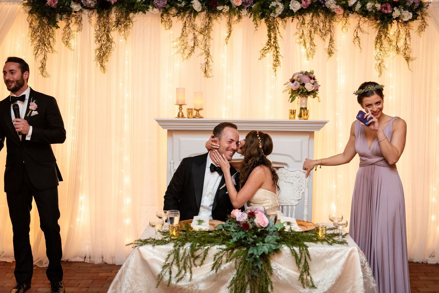 Bride and groom share a kiss at sweetheart table during wedding reception toast while bridesmaid reacts emotionally nearby.