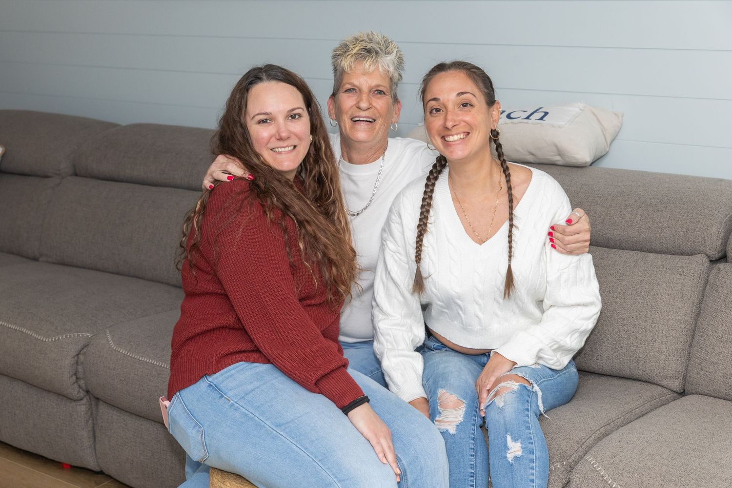 Three women smile together on a gray couch, two with long brown hair flanking an older woman in white.