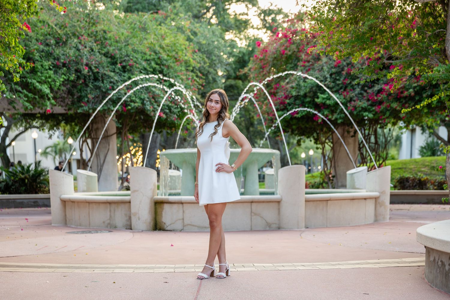 Young woman in white dress poses in front of decorative water fountain surrounded by lush green trees.