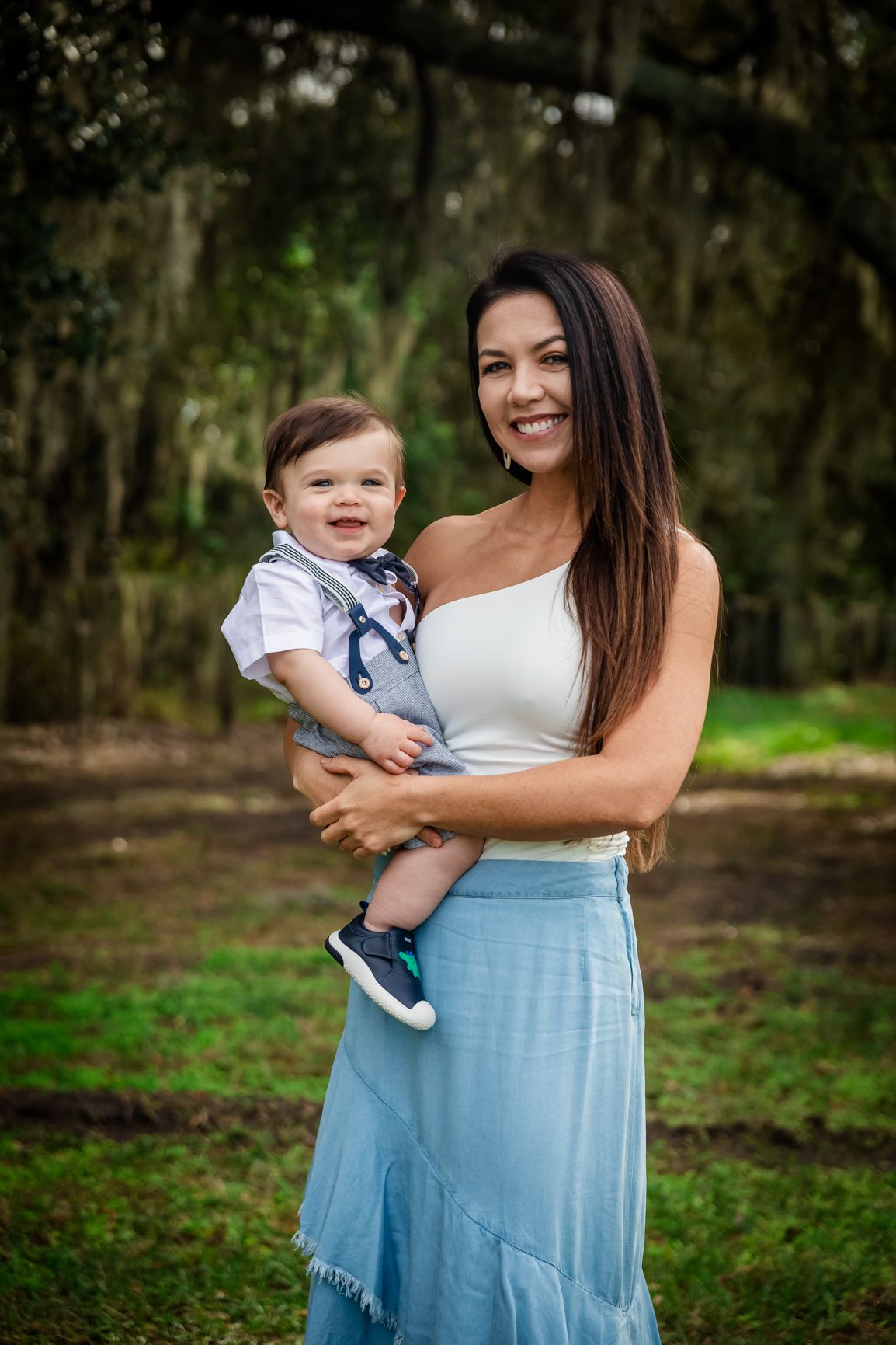 A smiling mother in a white top and blue skirt holds her baby boy outdoors among lush green trees.