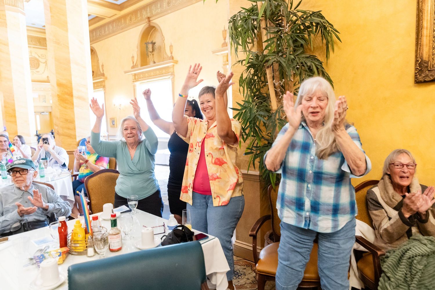 Elderly women clapping and raising hands in celebration at a Pride event in a decorated banquet hall.