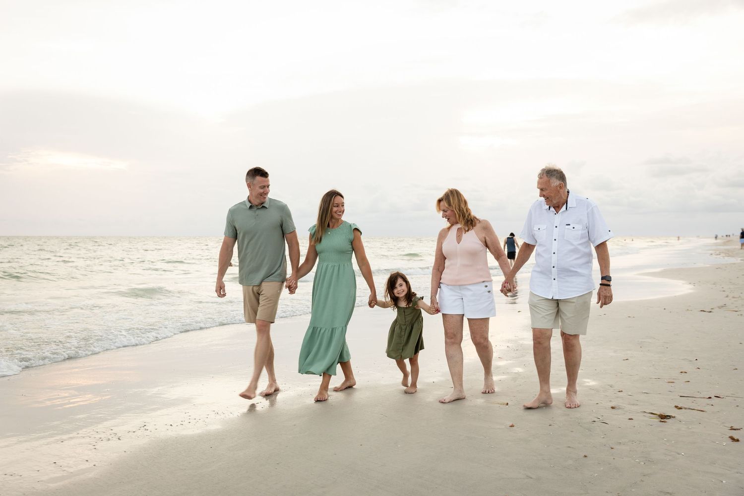 Family of five walking hand-in-hand along a sandy beach at sunset, wearing casual summer outfits.
