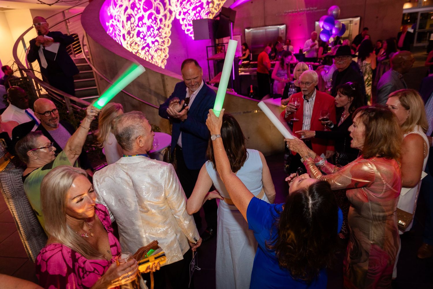 Guests dance and celebrate at a lively party, holding colorful glow sticks under vibrant pink lighting.