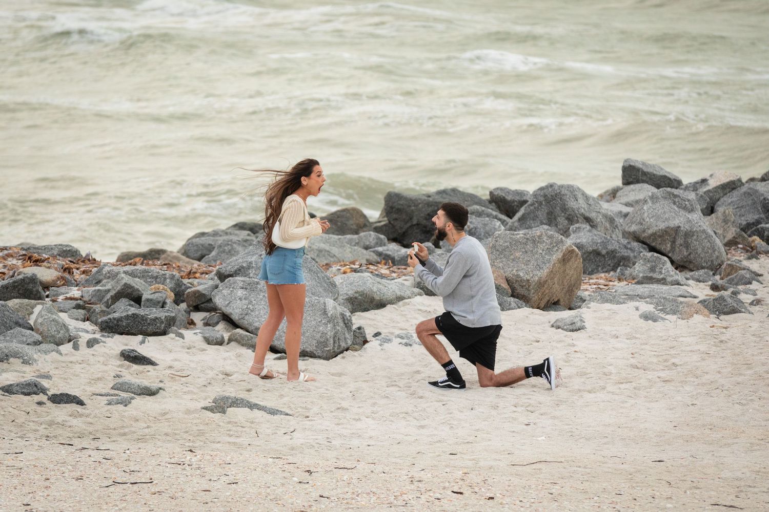 Man proposing on one knee to woman on a rocky beach shoreline.