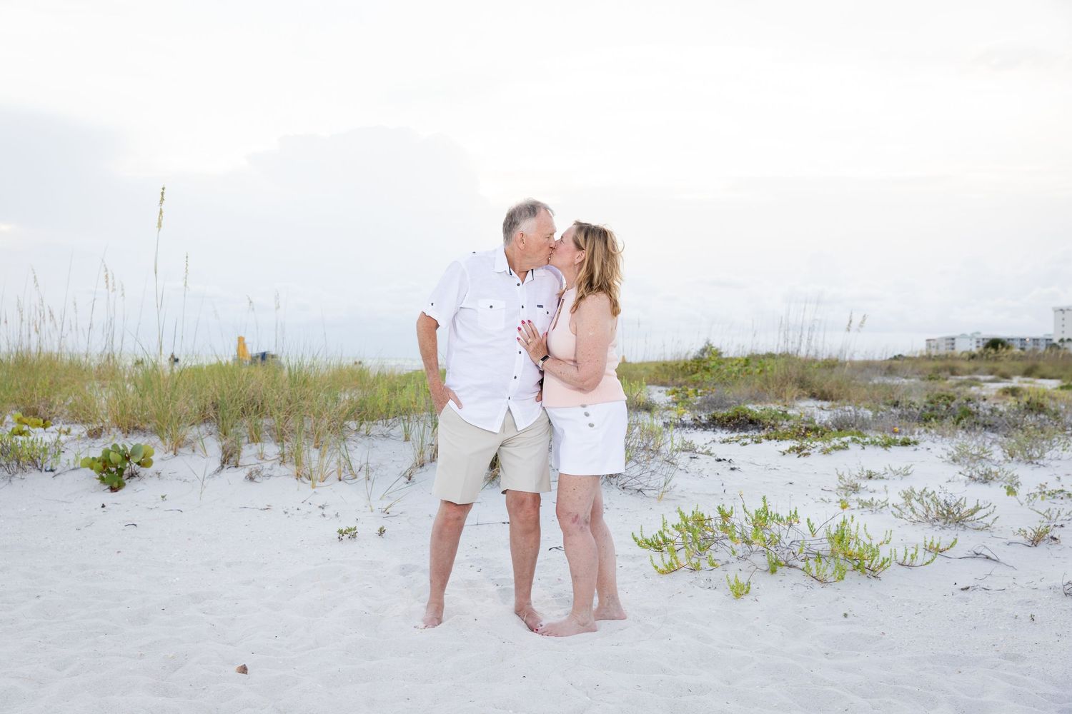 Couple sharing a romantic kiss on a white sandy beach with sea oats in the background.