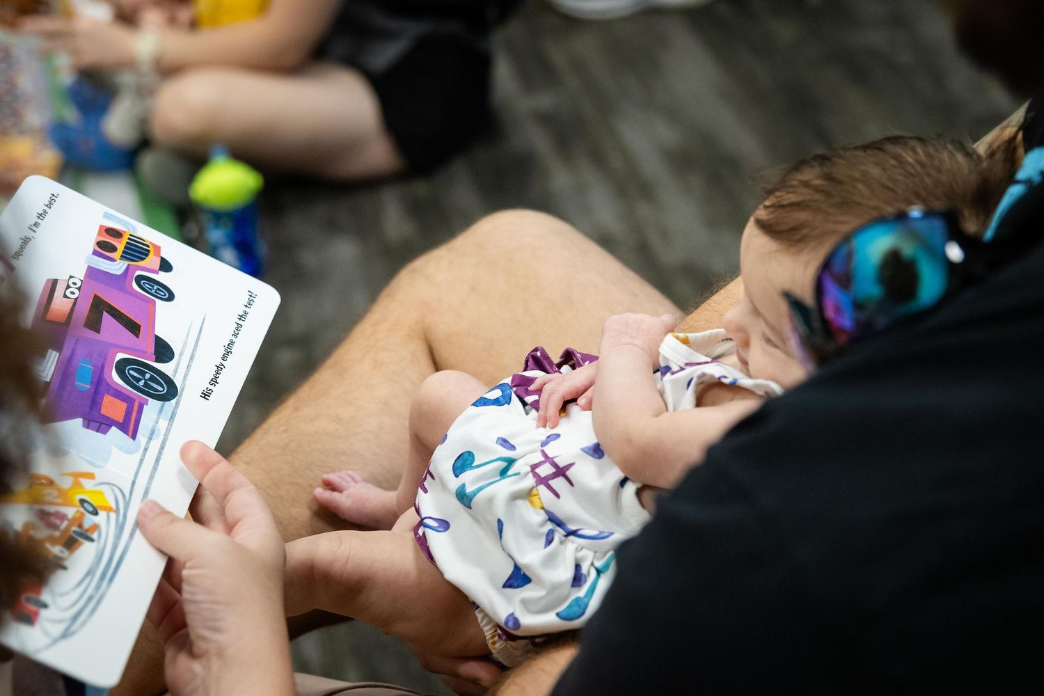 Adult reading a colorful children's book to a baby wearing a floral outfit and purple headphones.