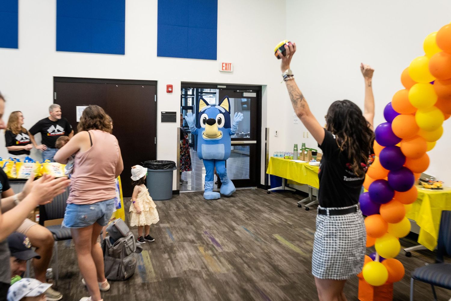 Blue mascot entertains guests at a festive party with purple and gold balloons and decorations in a bright room.