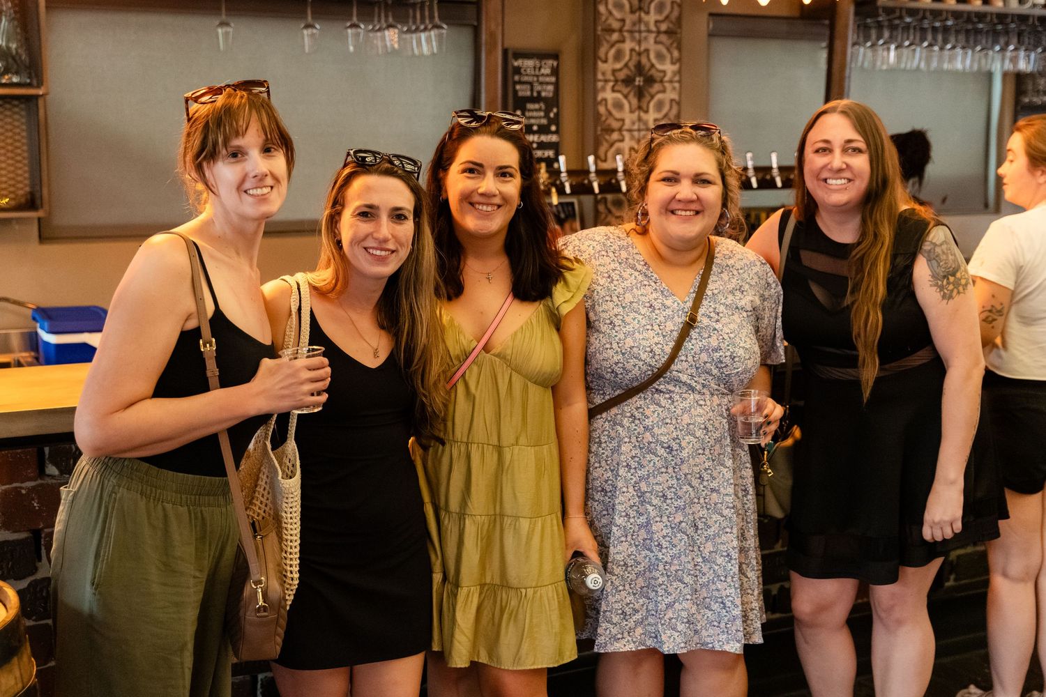 Five women smile together at a bar, dressed in summer outfits during a social event.