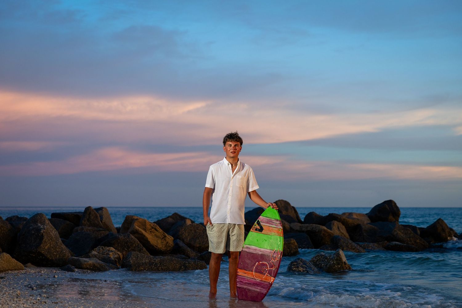 Young man holding a colorful bodyboard on a rocky beach at sunset with pink and blue sky.