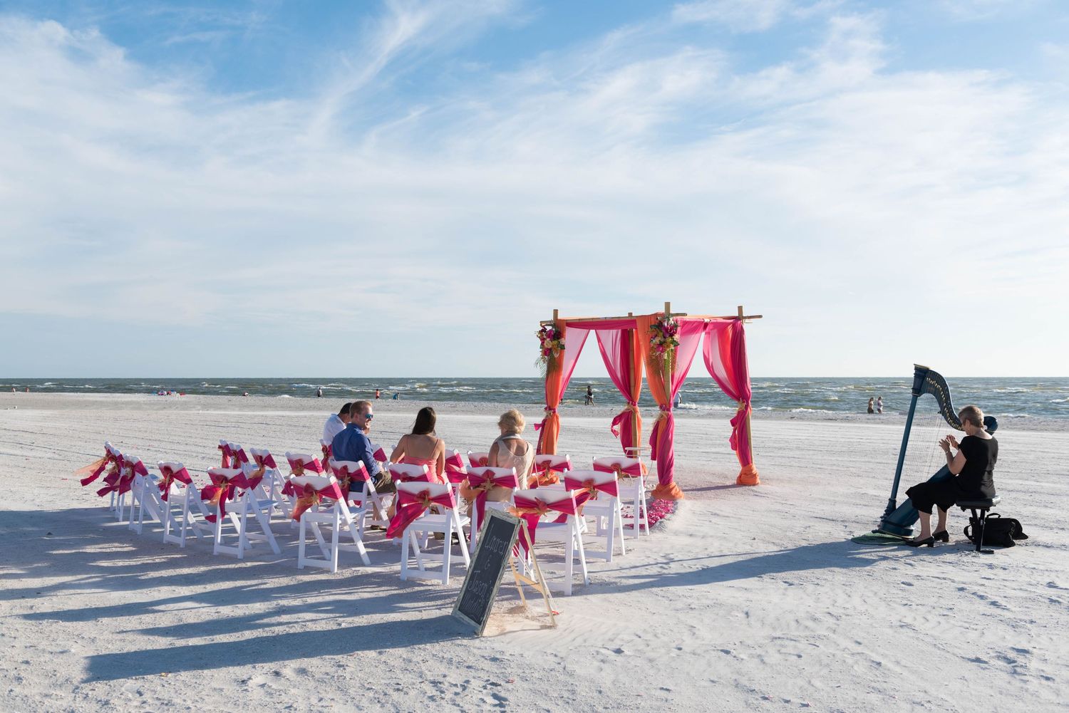 Colorful pink and gold beach wedding ceremony setup with harp player on white sand.