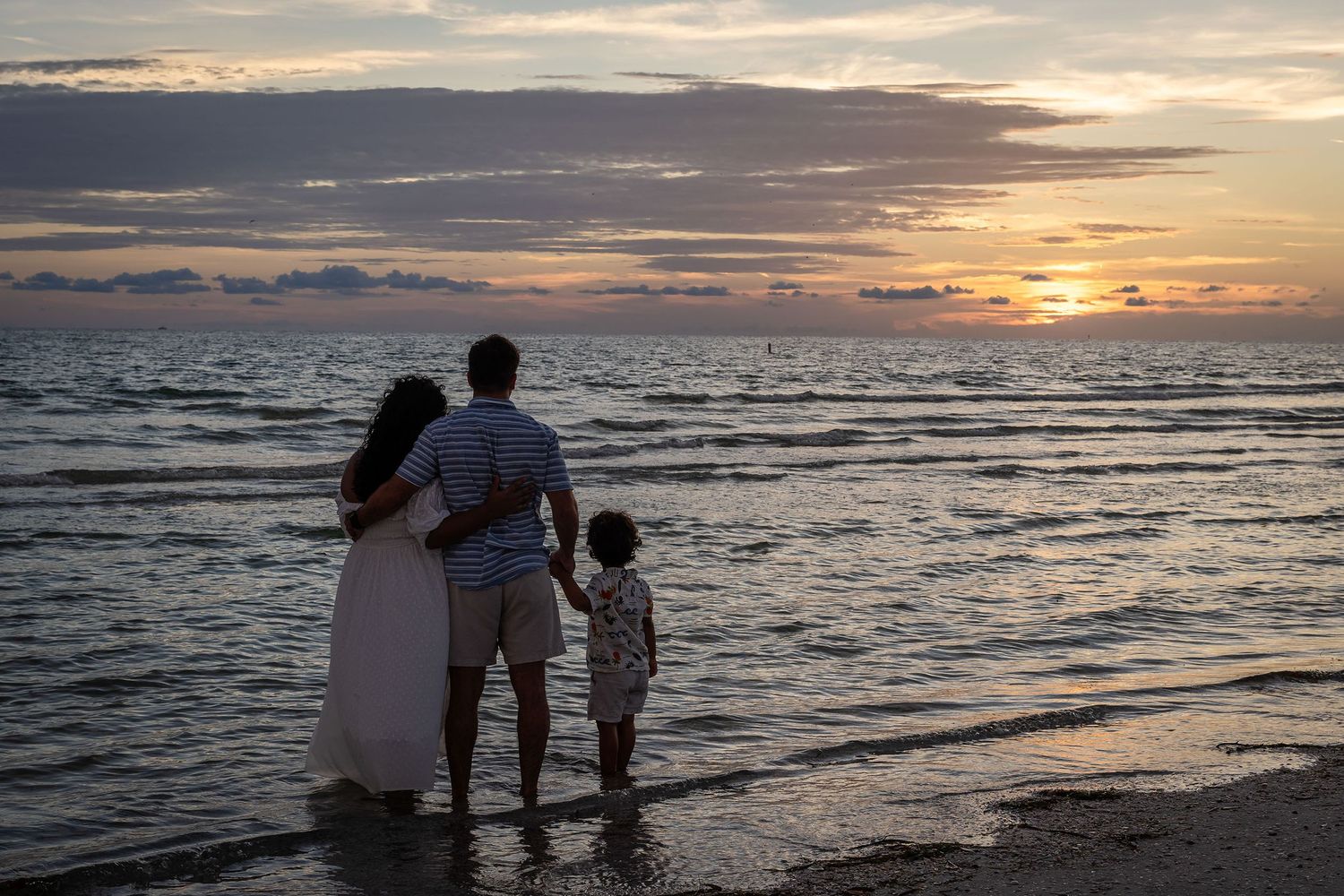 Family of three watching a beautiful sunset over the ocean waves on a serene beach at dusk.