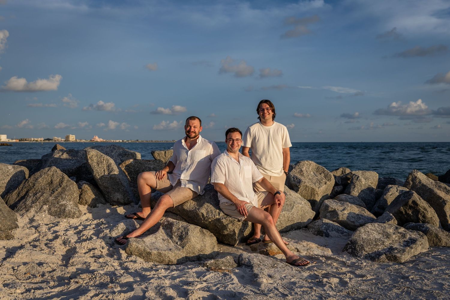 Three men posing on rocky beach shore with ocean waves and blue sky in background.