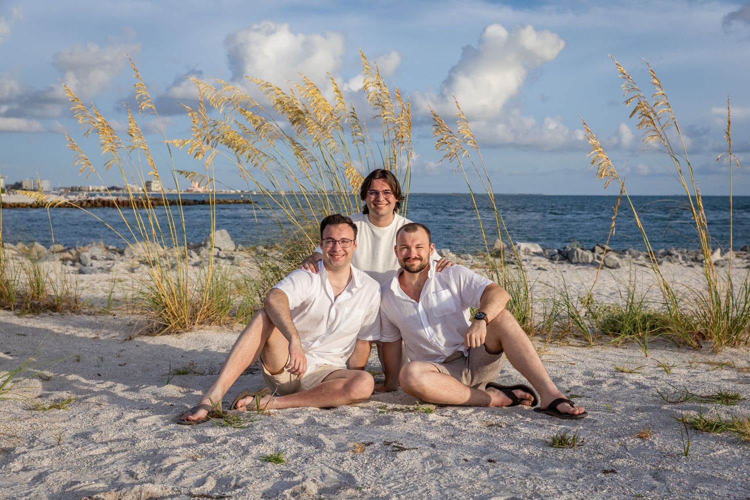 Three people smiling together on a sandy beach with sea oats and ocean in the background on a sunny day.