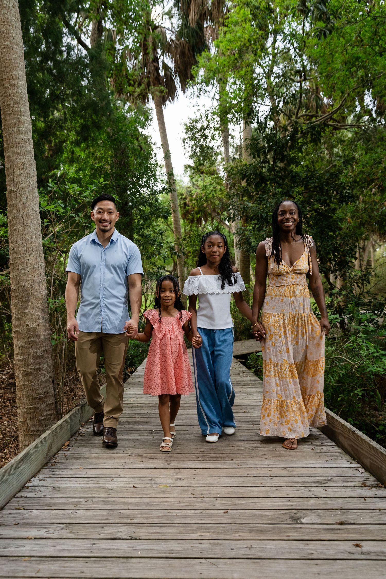 A joyful multiracial family of four walks along a wooden boardwalk surrounded by lush green trees.