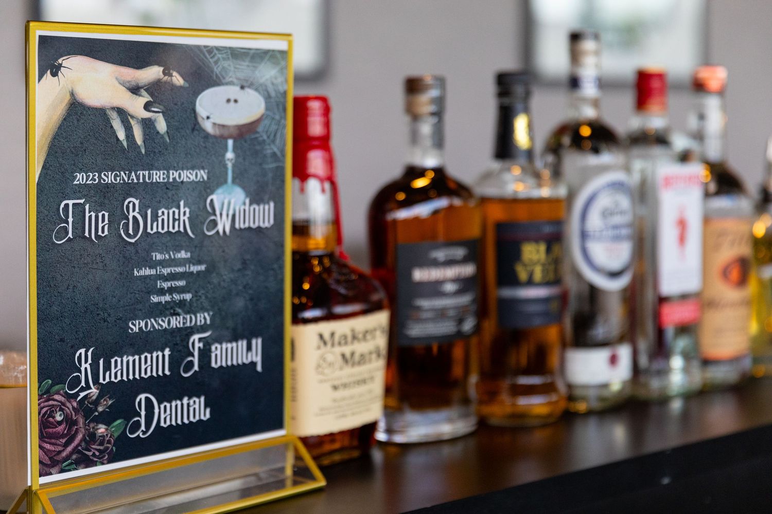A signature cocktail menu sign for The Black Widow drink sponsored by Klement Family Dental, displayed beside bottles of spirits.