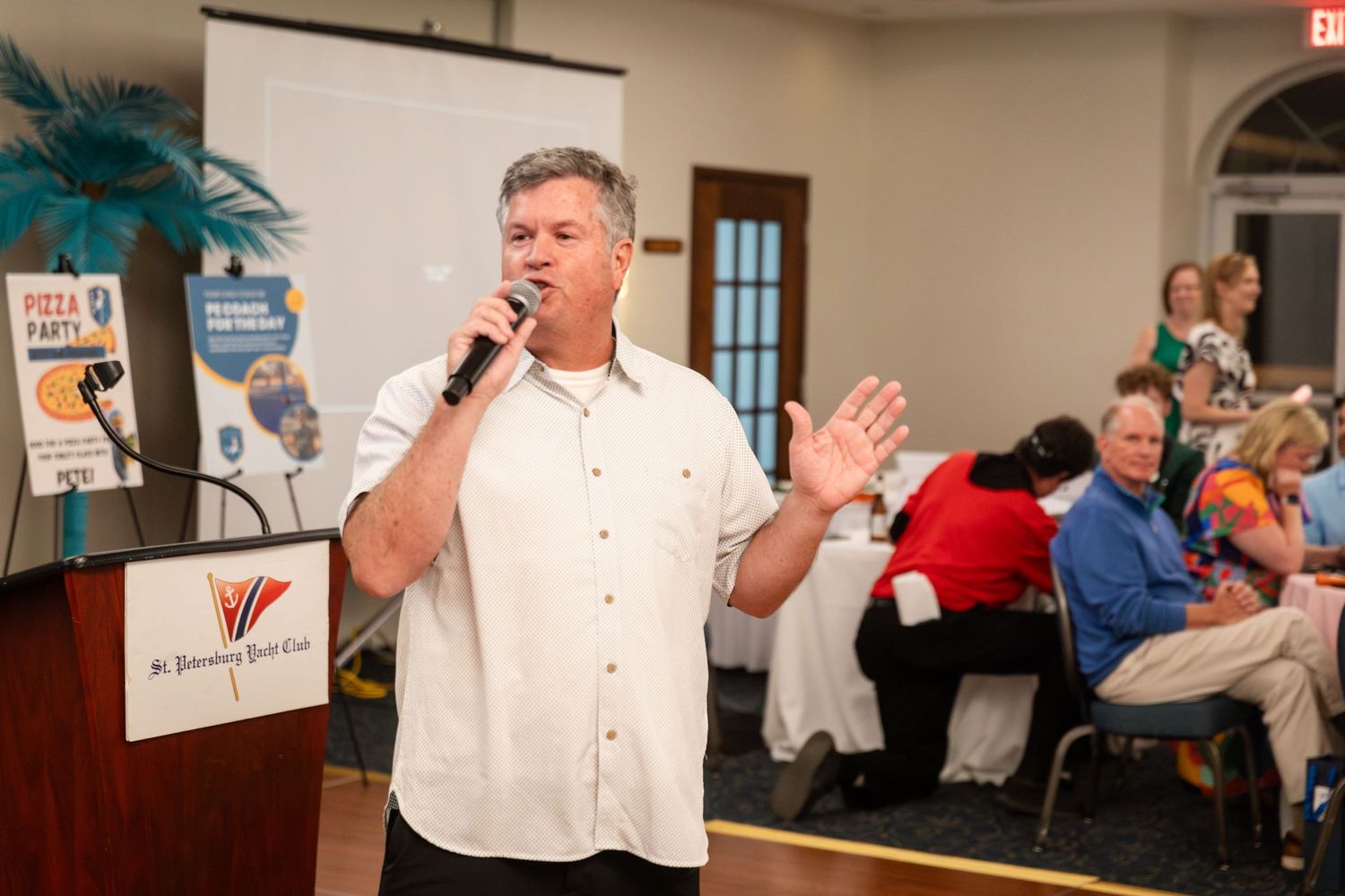 Man speaking into microphone at indoor event with audience seated at tables and presentation screen in background.