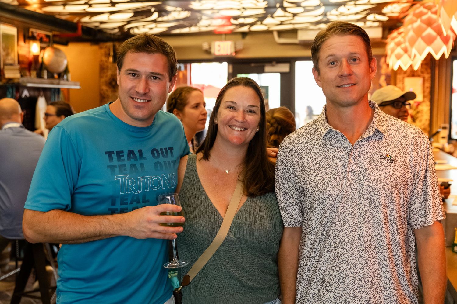 Three friends smiling and posing together at a casual indoor social event, one wearing a teal shirt.