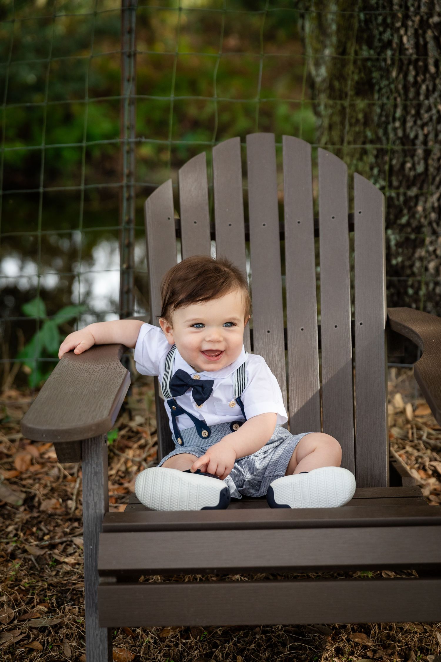 Smiling baby boy in bow tie and suspenders sitting in a wooden Adirondack chair outdoors.