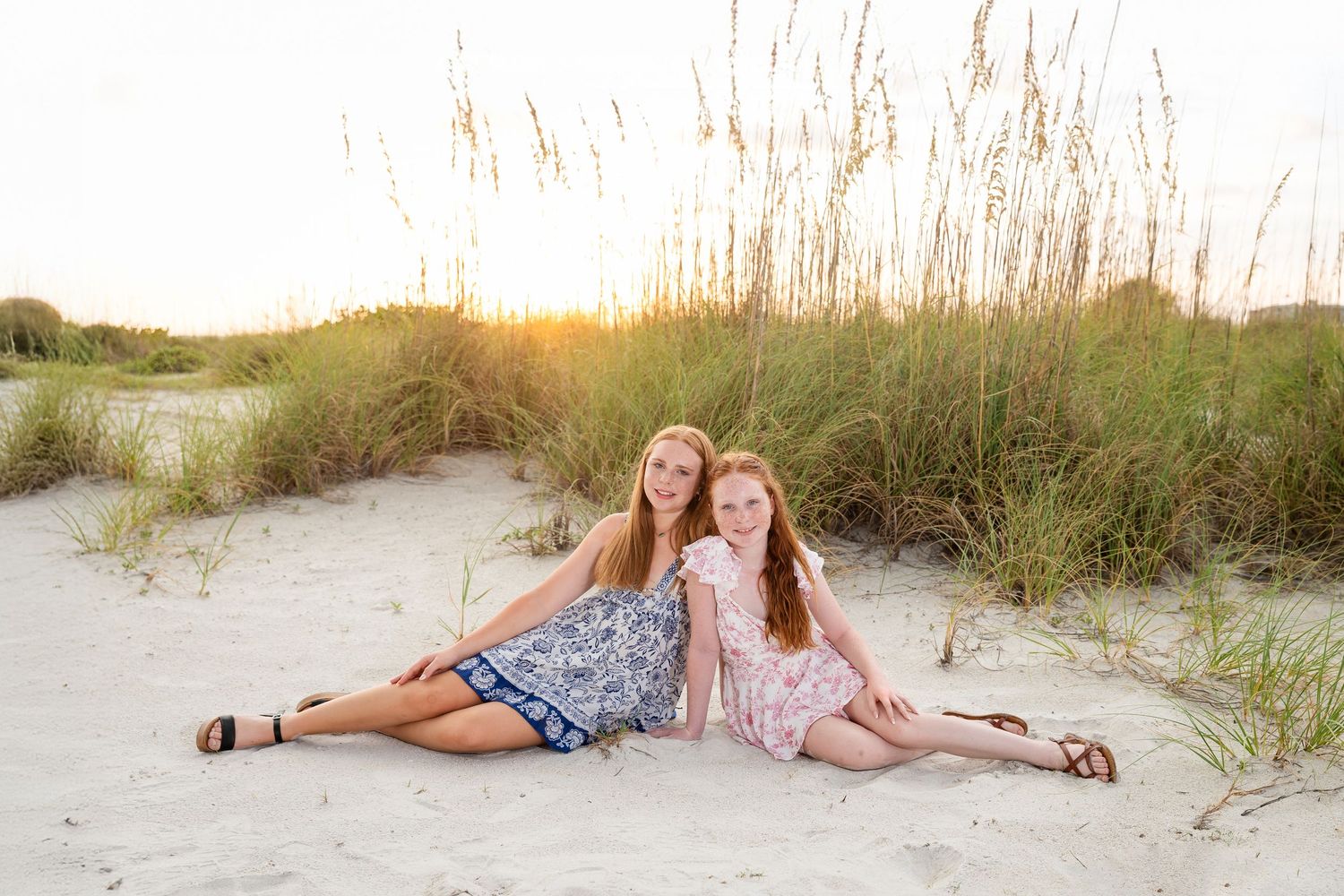 Two teenage girls sitting on sandy beach dunes at sunset, wearing floral dresses and sandals, smiling together.