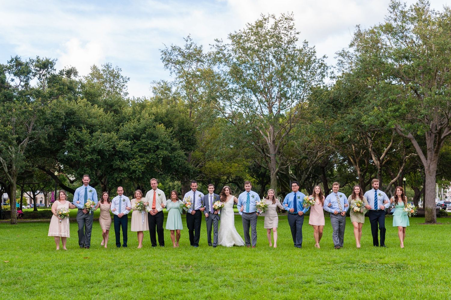 Large wedding party posing together on green grass with trees behind.
