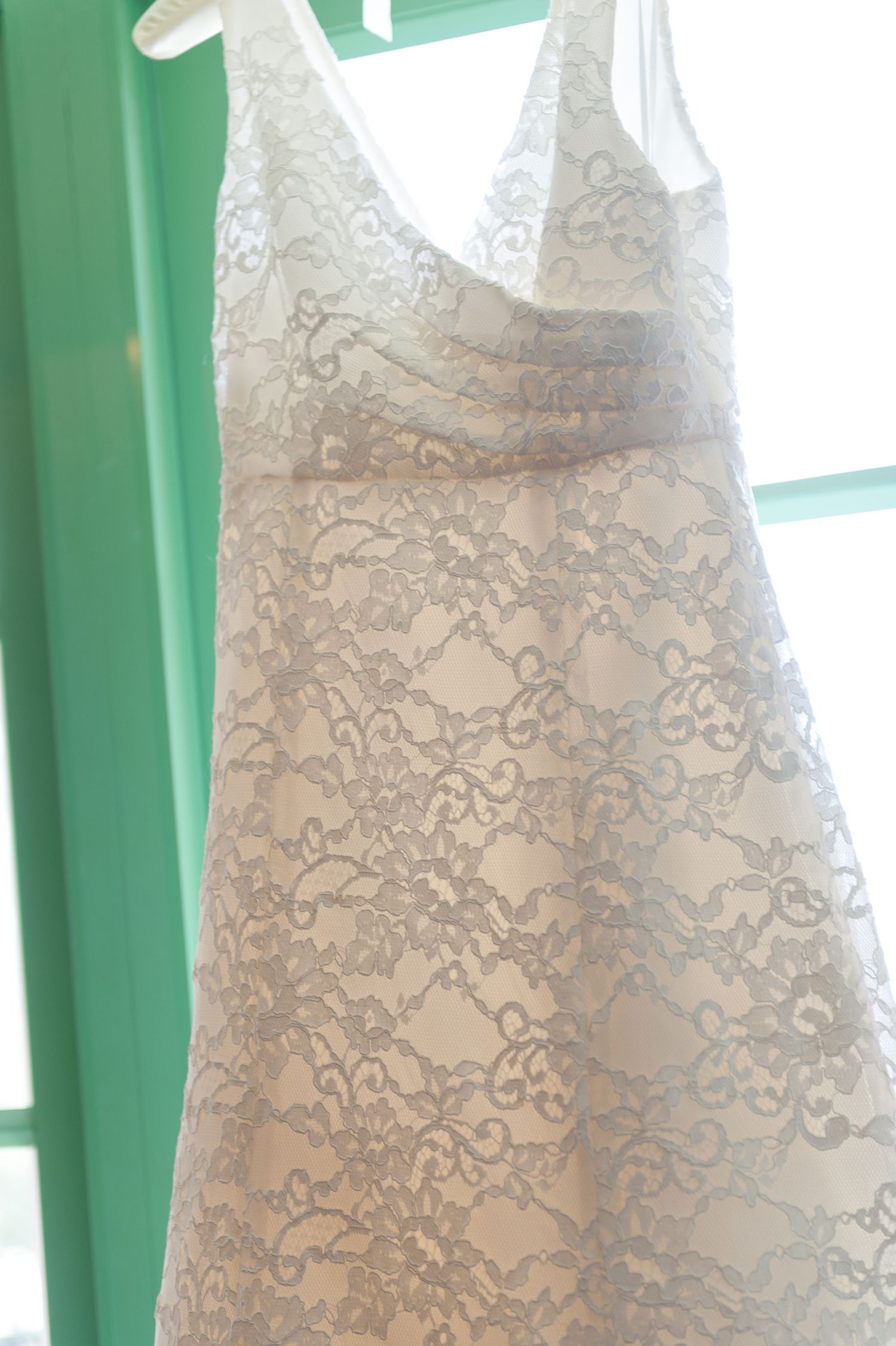 White lace wedding dress with silver embroidery and satin sash hanging on a hanger by a green window.