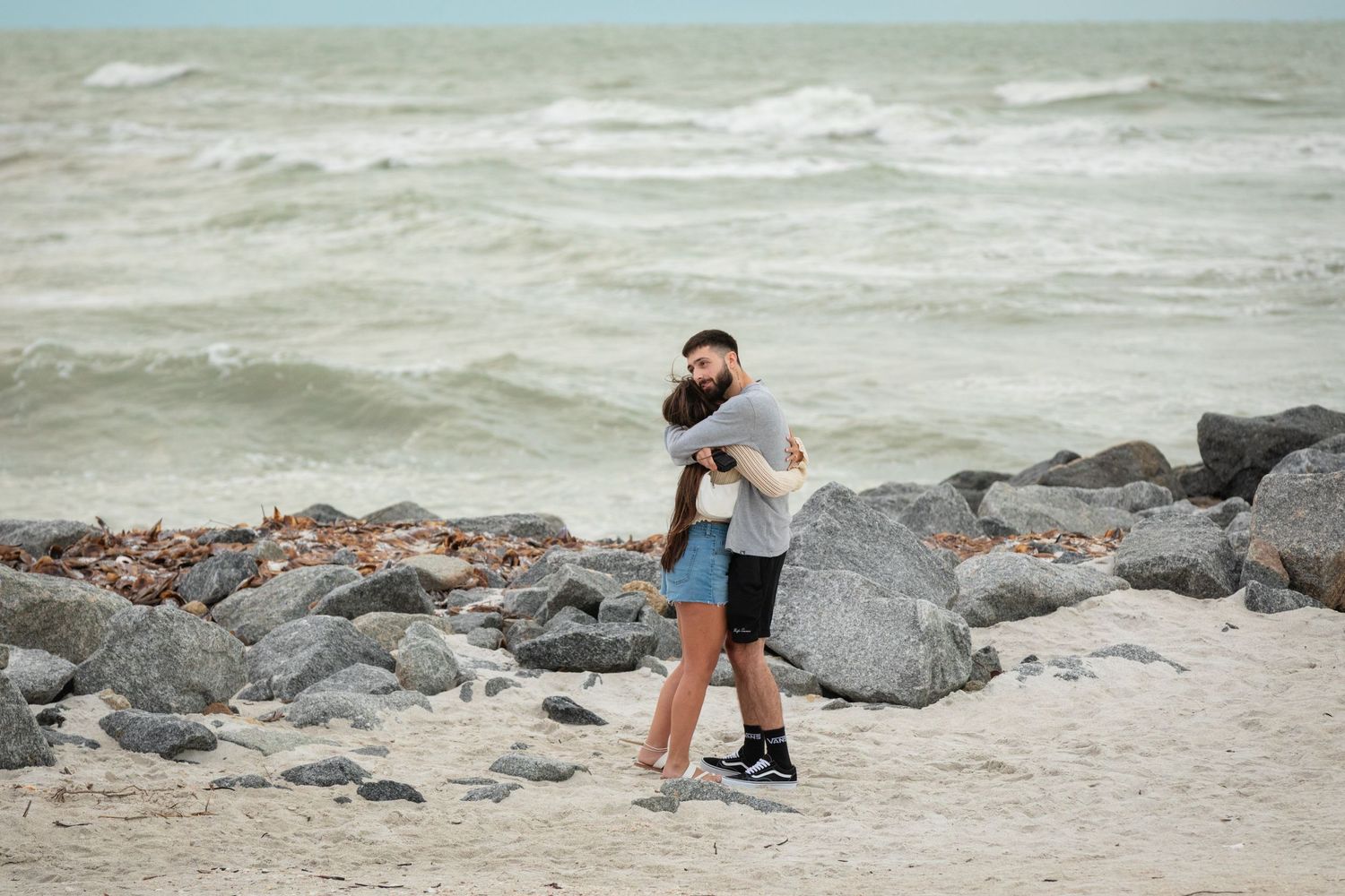 Couple embracing and hugging on a rocky beach with rough ocean waves in the background.