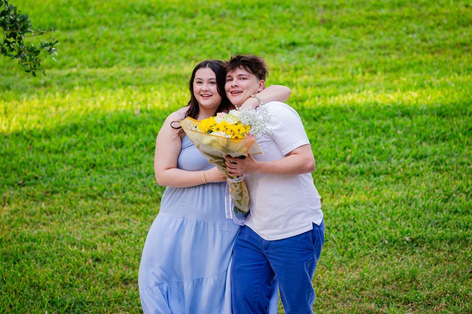 Couple posing outdoors on green grass, woman in blue dress and man in white shirt holding yellow flower bouquet.