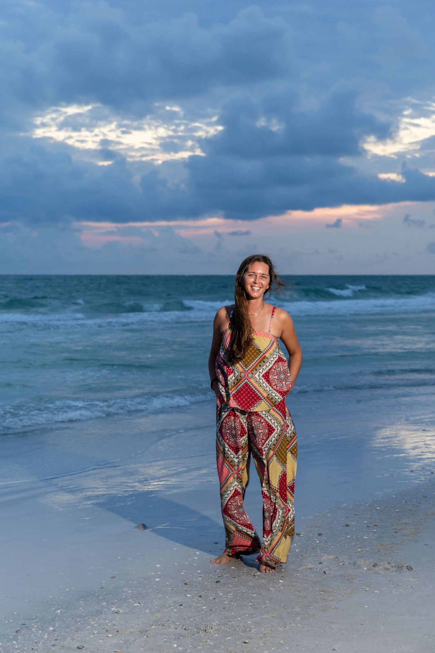Young woman in vibrant patterned jumpsuit smiling on a beach at sunset with ocean waves behind her.