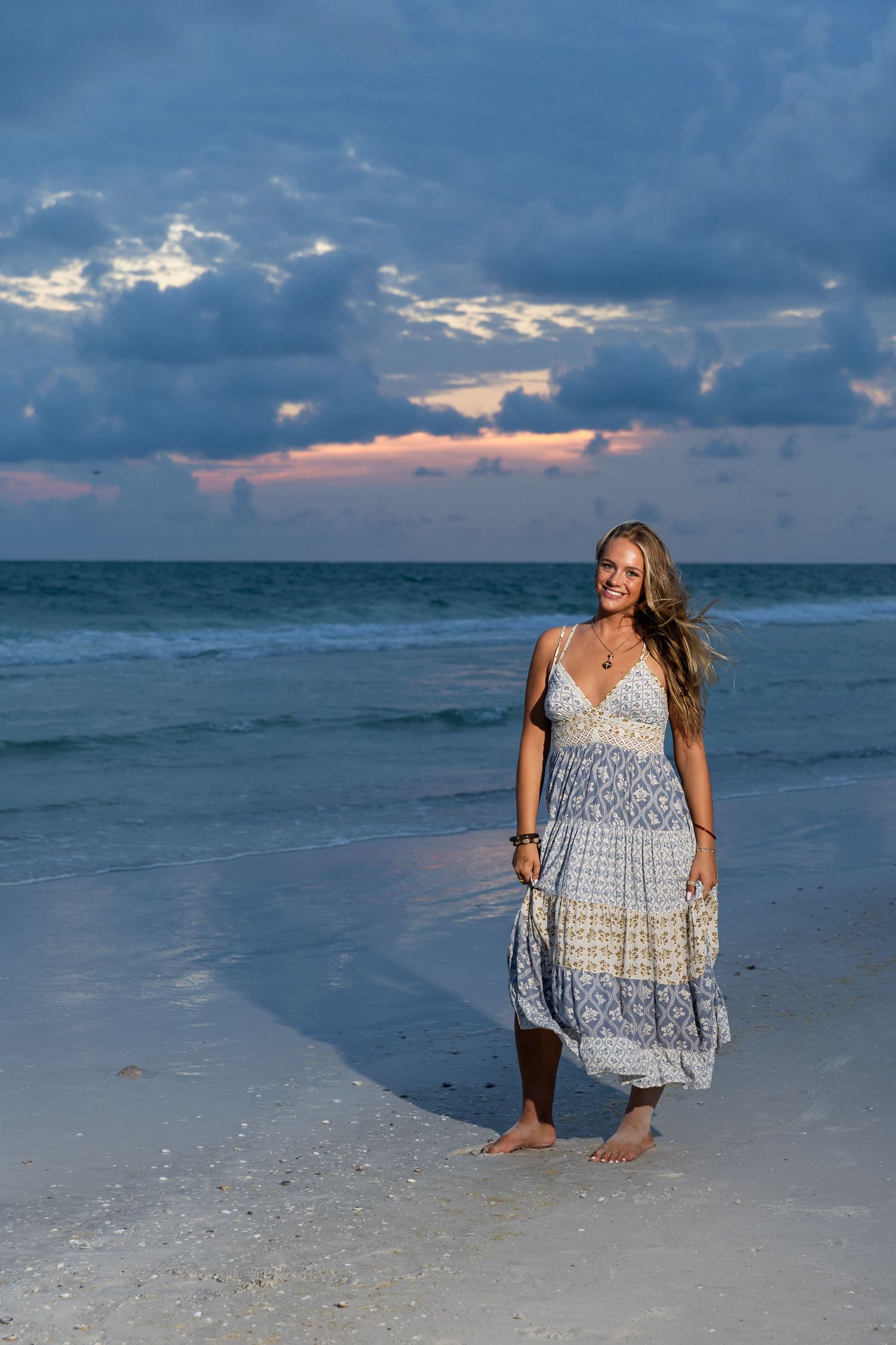 Woman in blue patterned sundress standing barefoot on beach shore during colorful sunset with ocean waves.