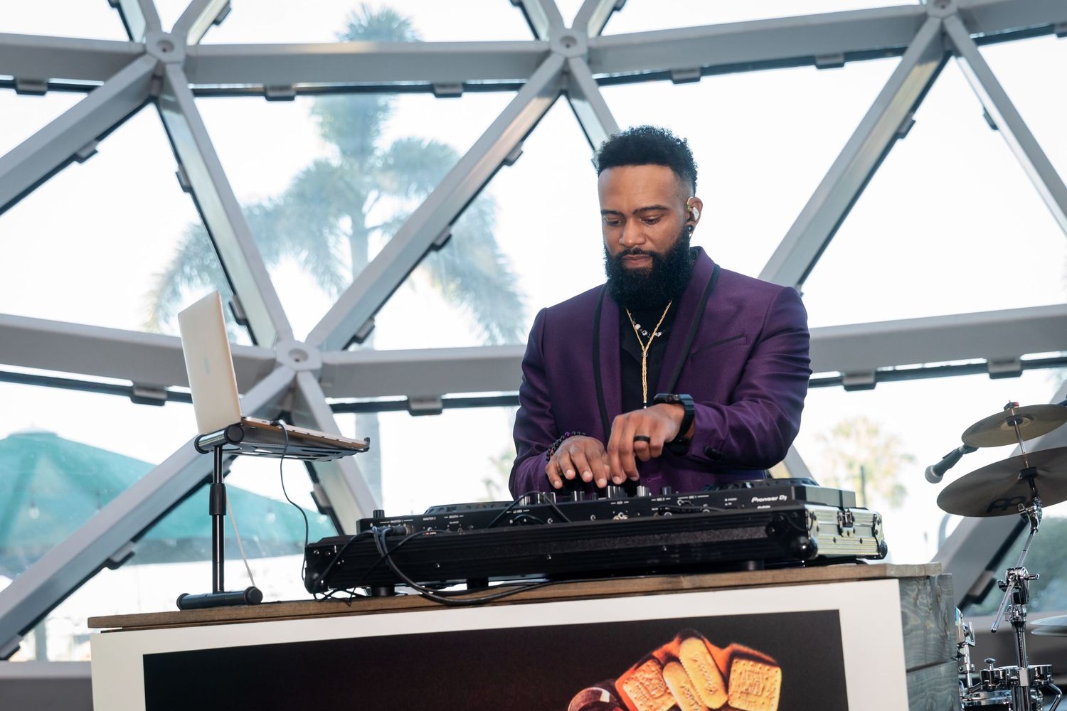 A DJ in a purple suit performs at a modern venue with a glass dome ceiling and drum kit in the background.
