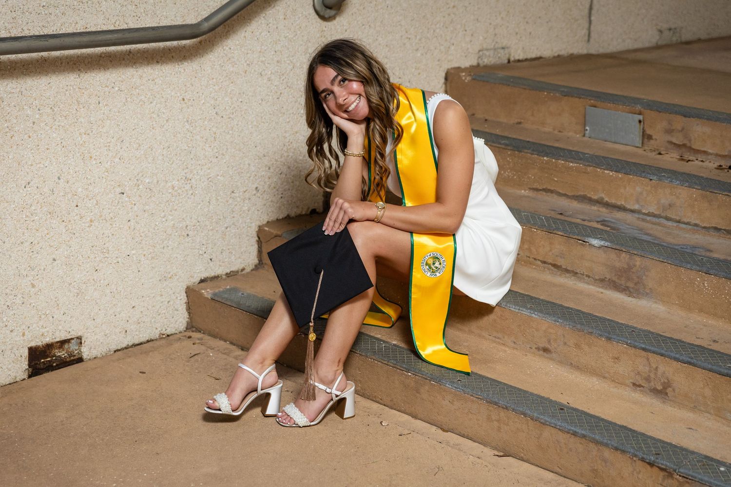 Smiling graduate in white dress and yellow sash sitting on concrete steps holding a diploma.