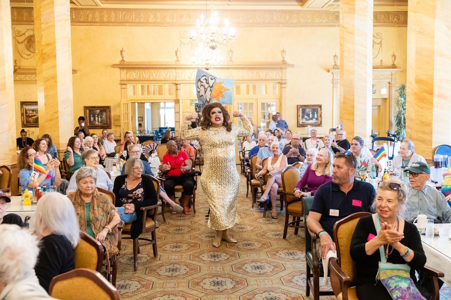 A drag performer in a gold dress walks the aisle at a Pride celebration in a grand ballroom with cheering crowd.