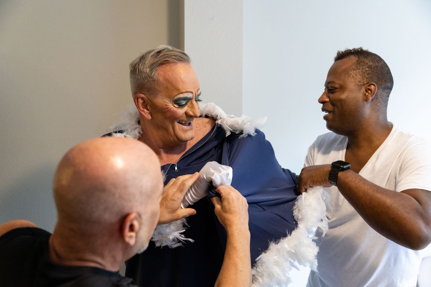 Three men laughing together, one wearing a blue outfit with a feather boa in a fun, candid indoor moment.