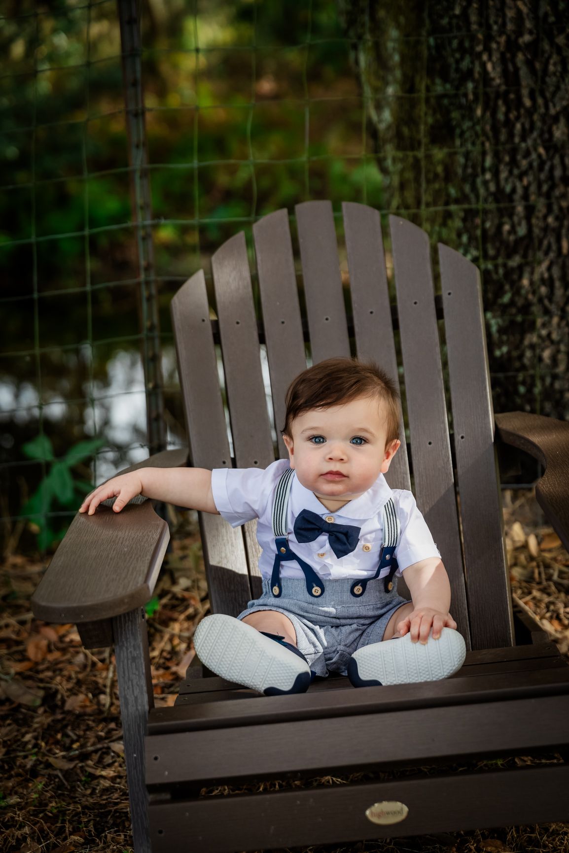 Cute infant boy wearing suspenders and bow tie posed in a brown Adirondack chair in a garden setting.