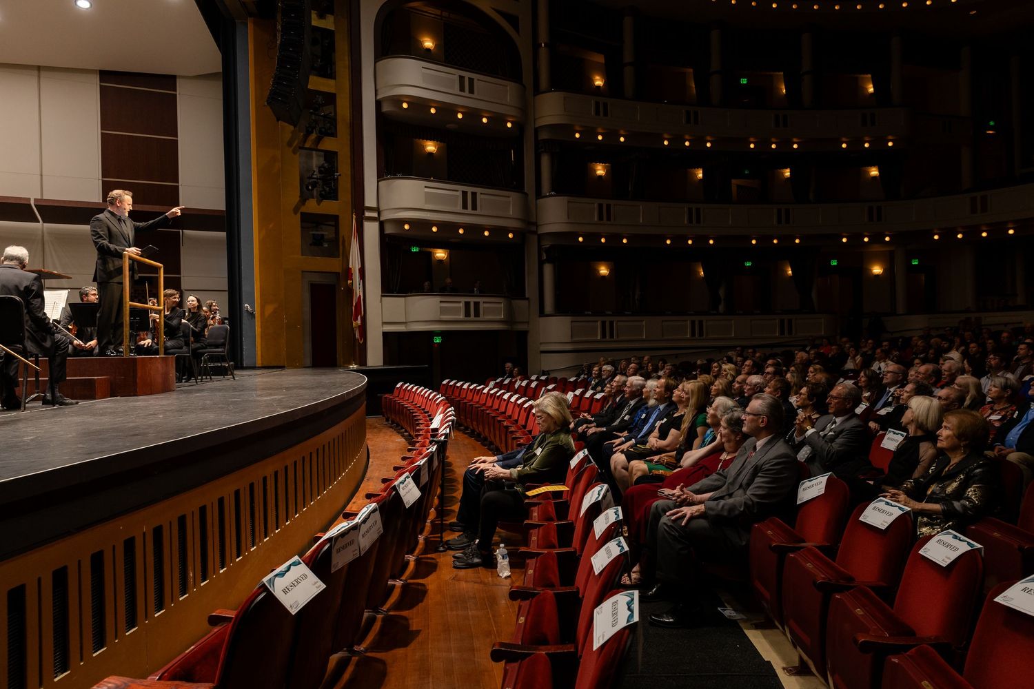 Conductor leads orchestra performance before a large seated audience in an elegant theater.