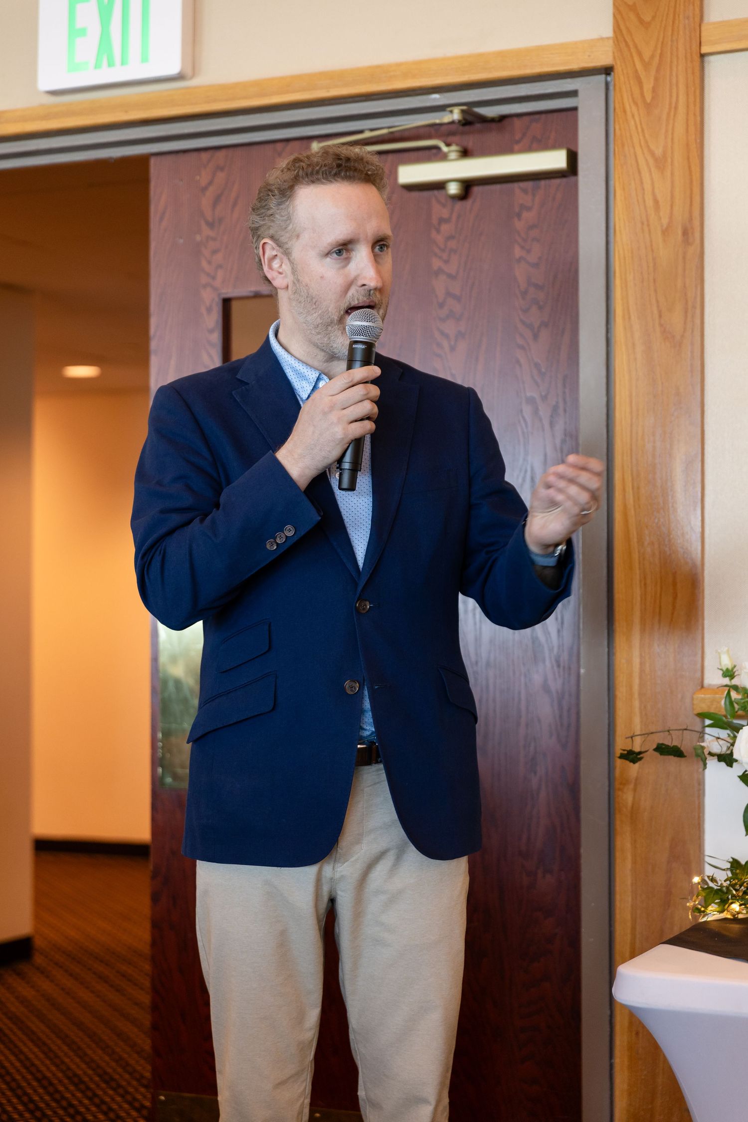 Man in navy blazer speaks into microphone at indoor event near exit sign.