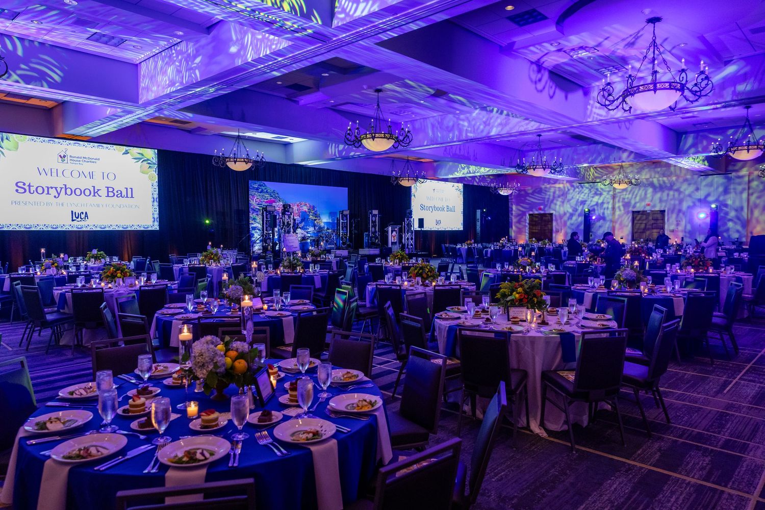 Elegant Storybook Ball gala dinner with round tables, blue lighting, floral centerpieces, and large screens in a grand ballroom.