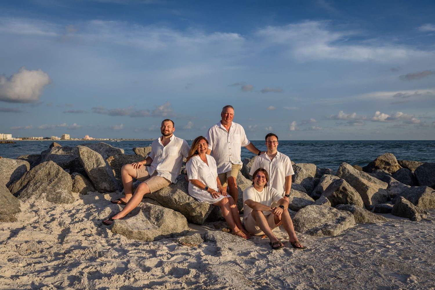 Family of five posing on rocky beach with ocean backdrop under blue skies in matching white outfits.