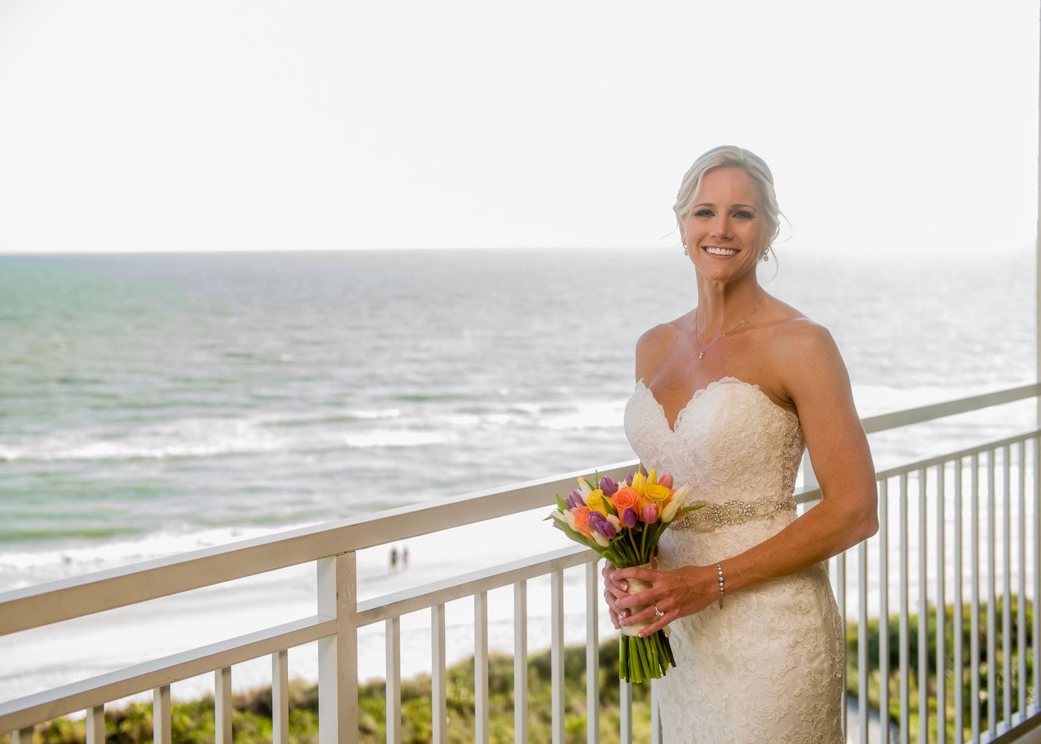 Smiling bride in strapless lace gown holding colorful bouquet on oceanfront balcony with blue sea backdrop.