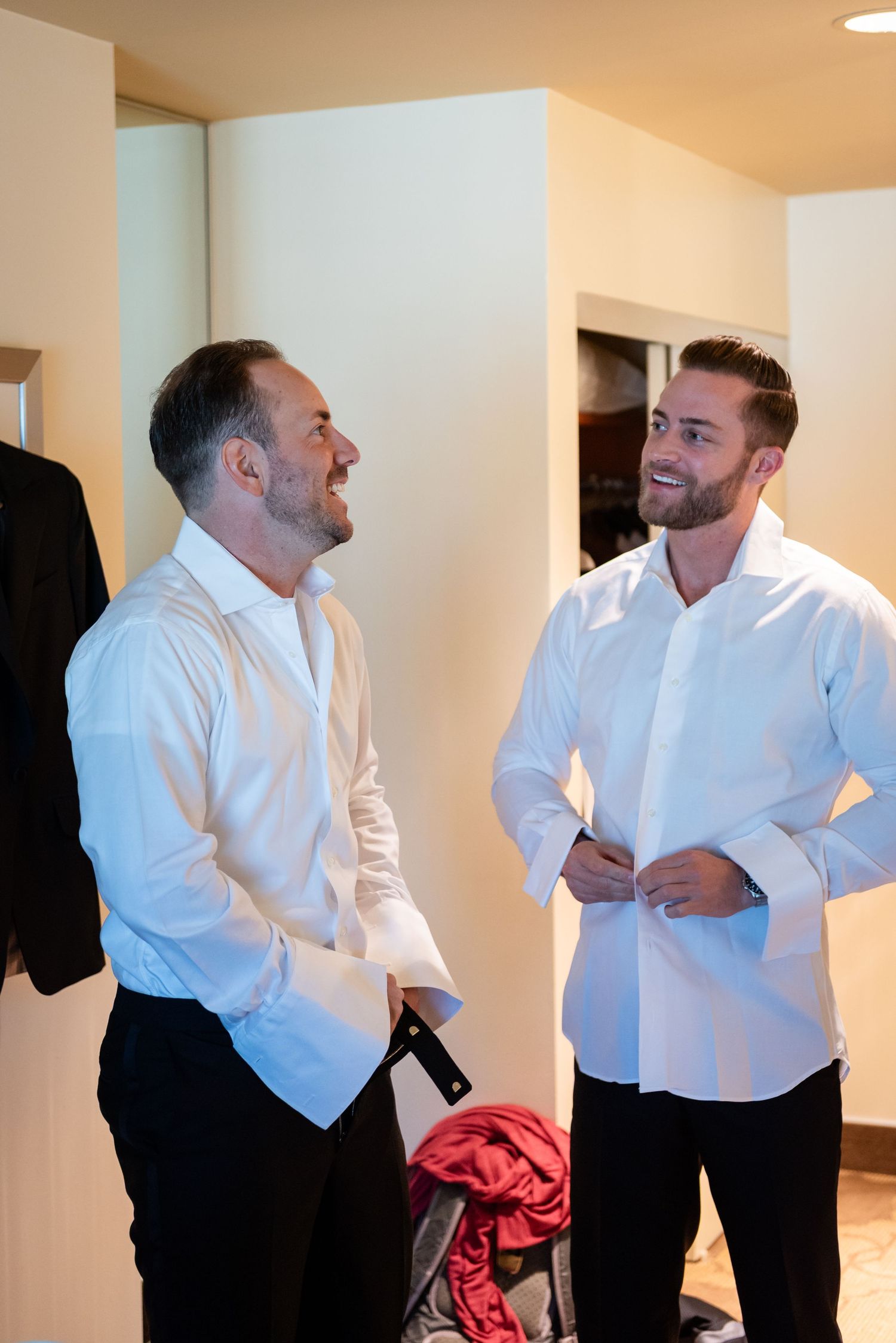 Two men in white dress shirts laughing together while getting ready for a wedding.