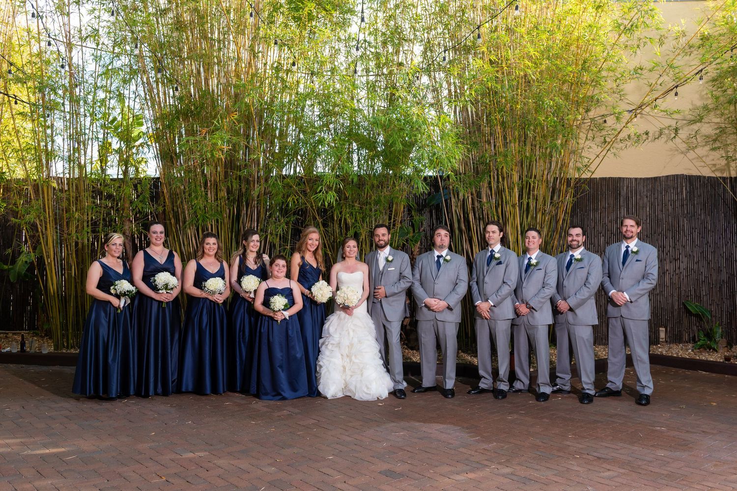 Wedding party in navy blue gowns and gray suits posing outdoors near bamboo trees.