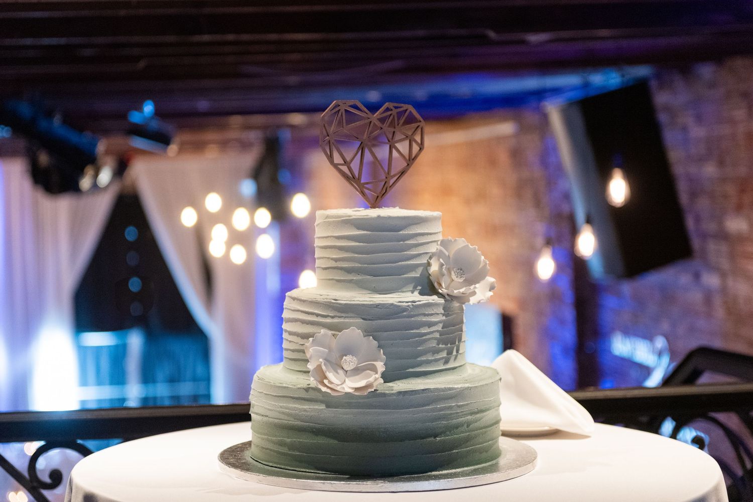 Three-tier ombre blue wedding cake with geometric heart topper and floral accents on a white table in an elegant venue.